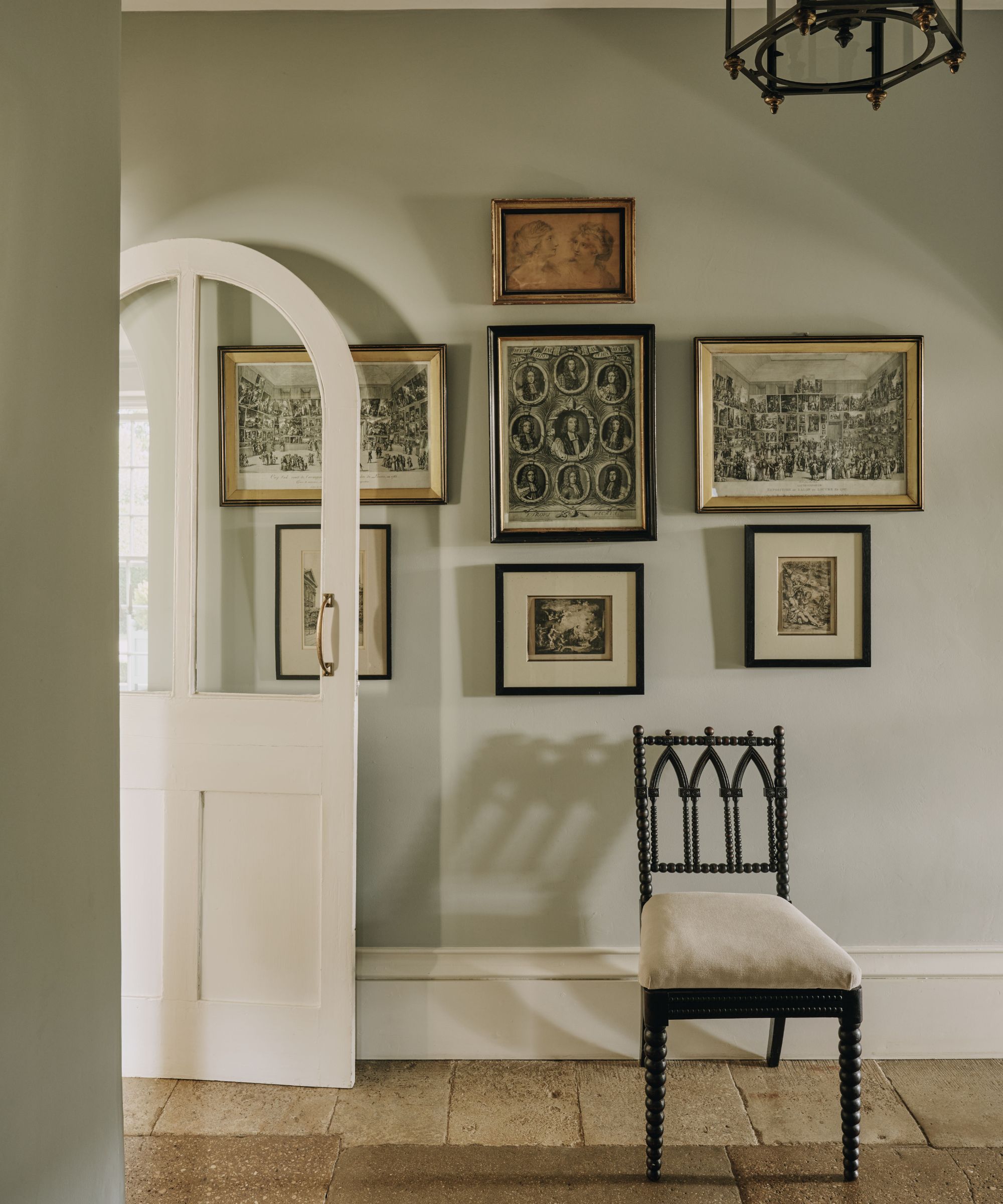 Hallway featuring a gallery wall of framed vintage sketches, a white arched door, stone floors, and a black bobbin-leg chair