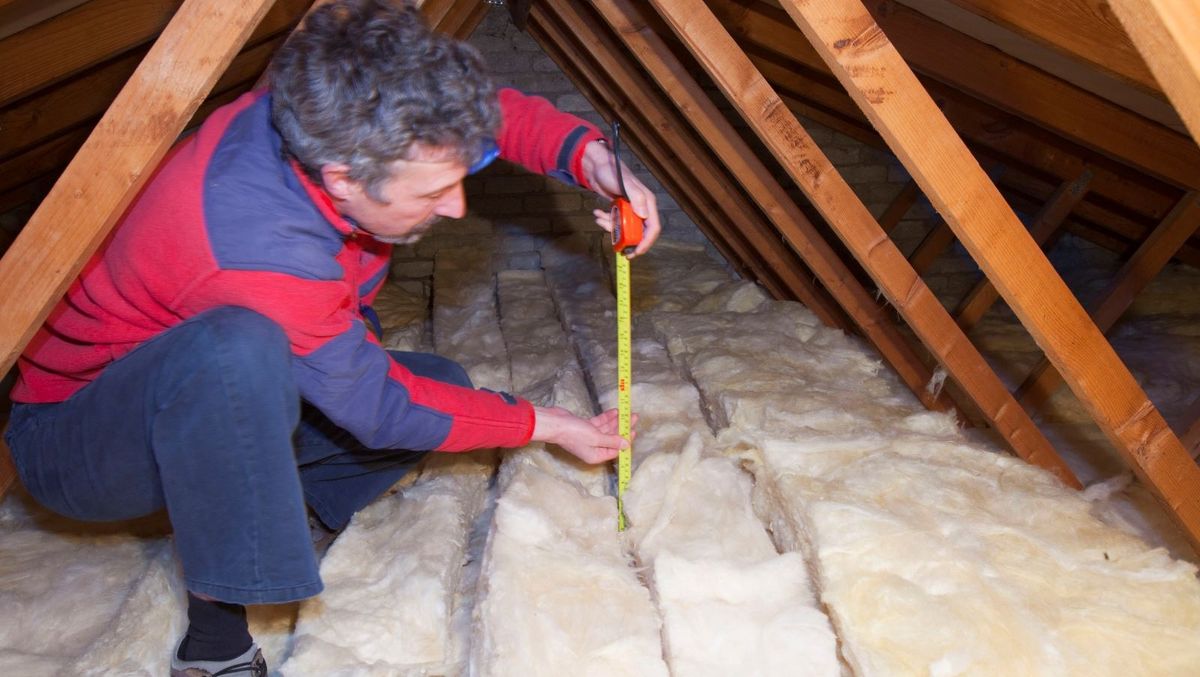 A man in a red top uses a tape measure to measure the thickness of loft insulation