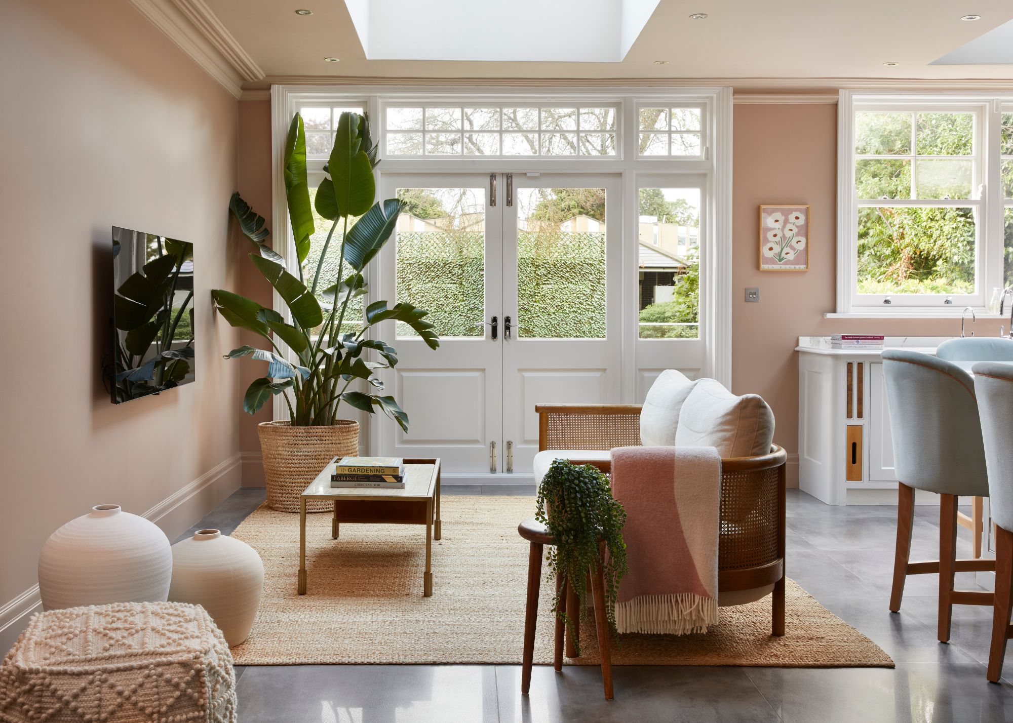 Open-plan kitchen and living room with pale pink walls, French doors leading into the garden, and a sofa opposite a TV mounted on the wall.