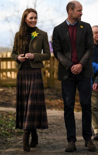 Kate Middleton wearing a long plaid skirt and cropped jacket with a daffodil pin walking on a sidewalk with Prince William, also wearing a daffodil pin