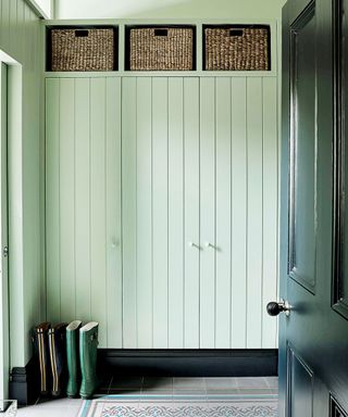 Light green painted tongue and groove paneling with storage cupboards, dark green painted front door, three woven storage baskets, stone flooring with patterned rug and two pairs of wellington boots