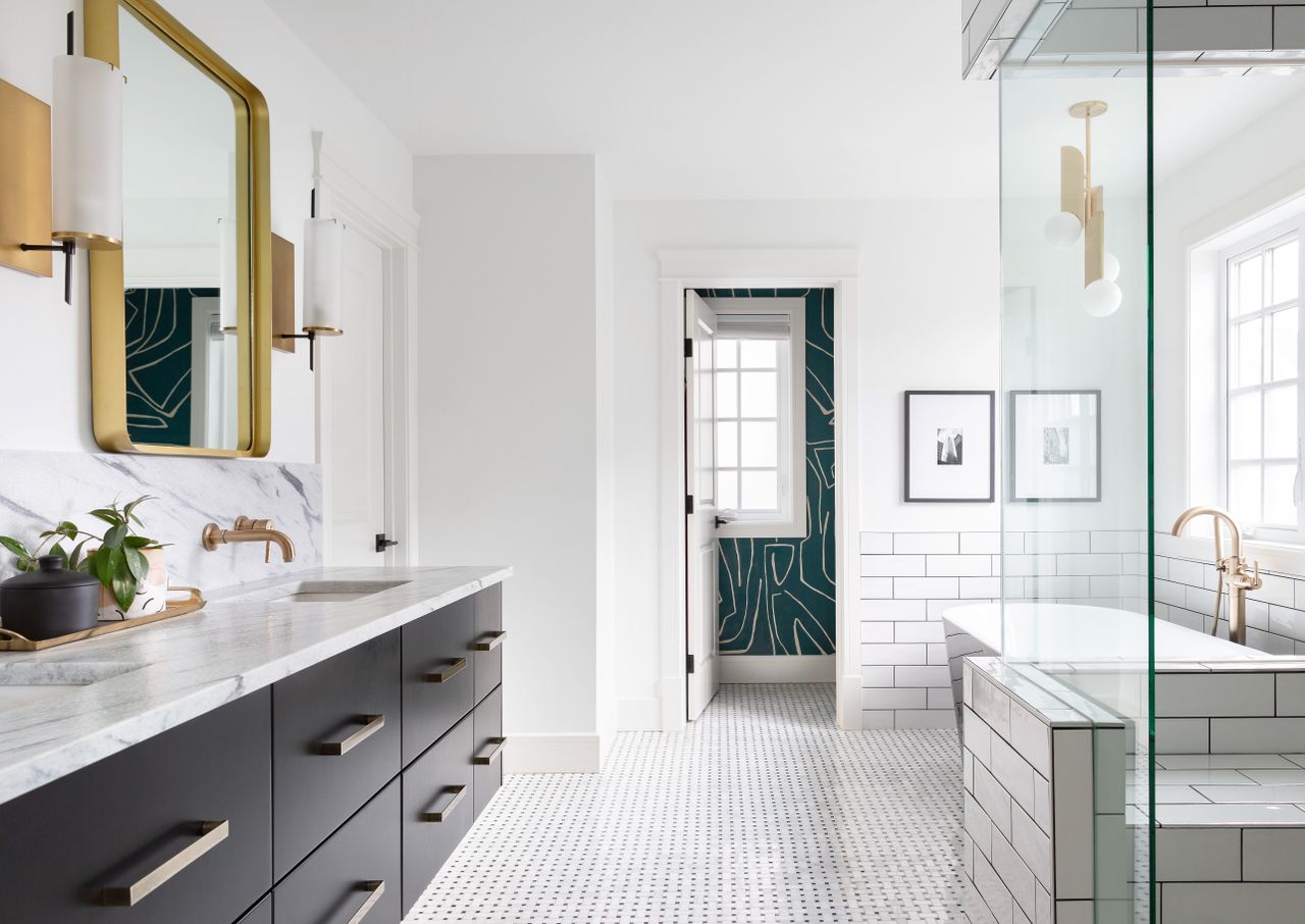 An all white bathroom with a mirror and lights in gold, and the toilet section with a wallpaper