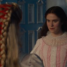 margot robbie as cathy with a corseted braid in her hair from the back looking at alison oliver as isabella waring a pink gown with bows in wuthering heights