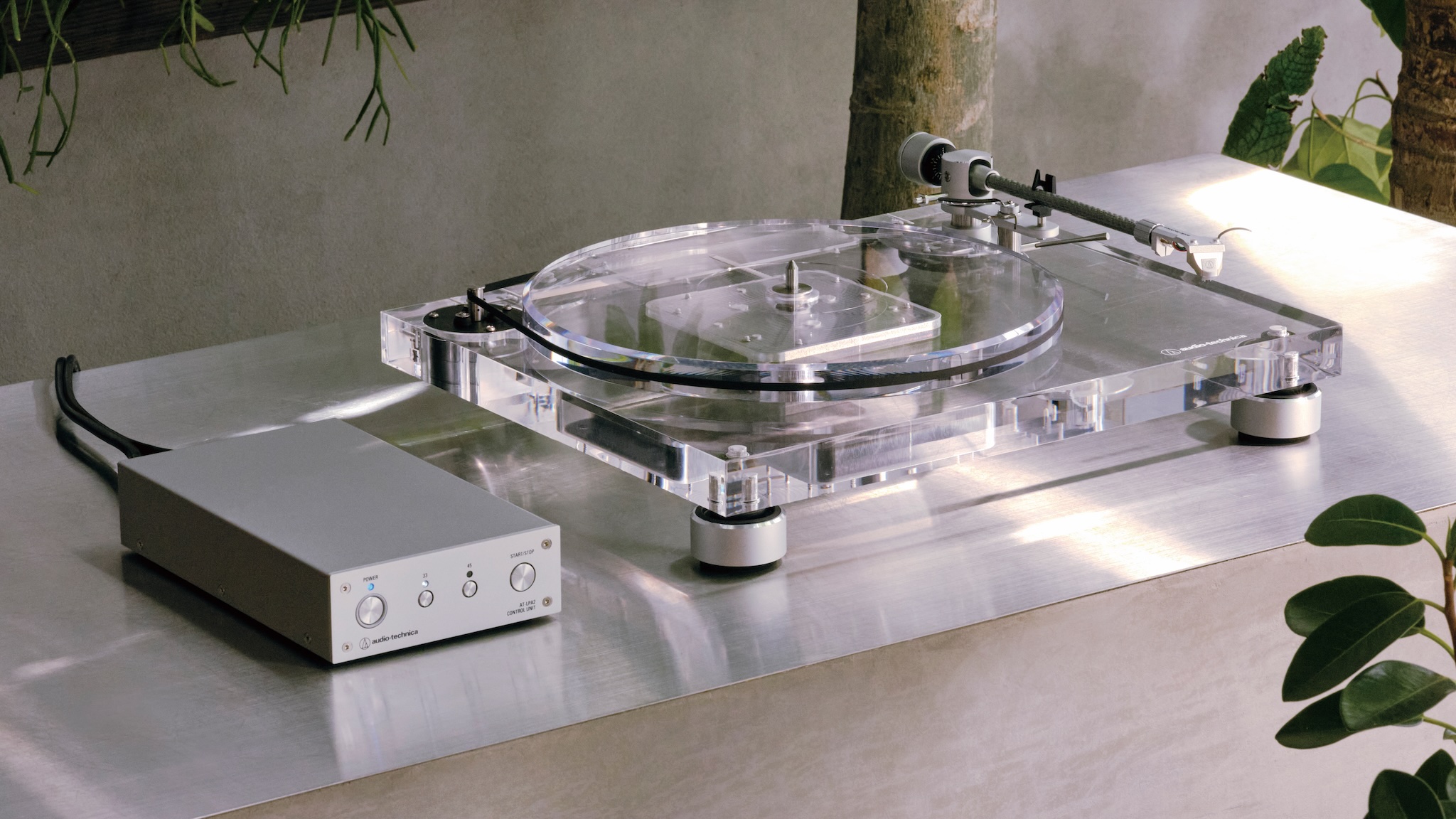 A clear acrylic Audio-Technica AT-LPA2 turntable on a shiny metal surface in a brightly lit room. It is conencted to a compact silver-coloured control box.