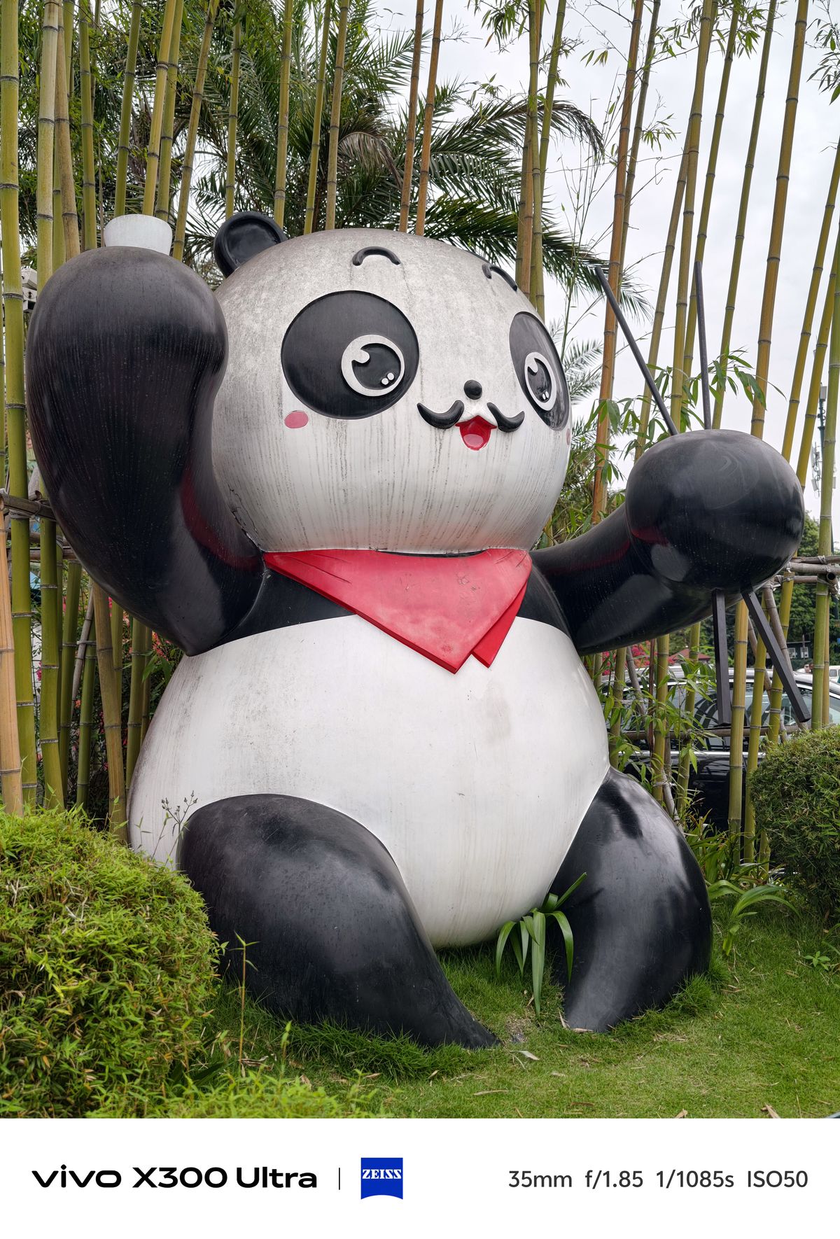 A large, cheerful cartoon panda statue wearing a red scarf, positioned in a grassy area in front of a tall thicket of green bamboo stalks.