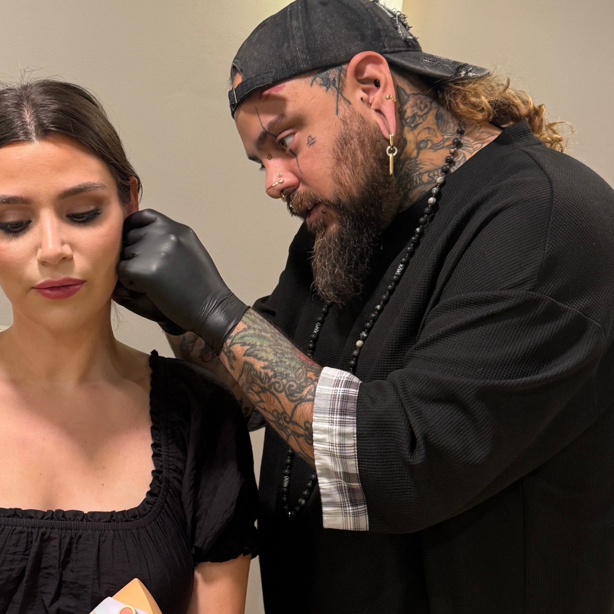 A girl sits in a chair while a tattooed piercer wearing gloves pierces their ear in a jewelry store.