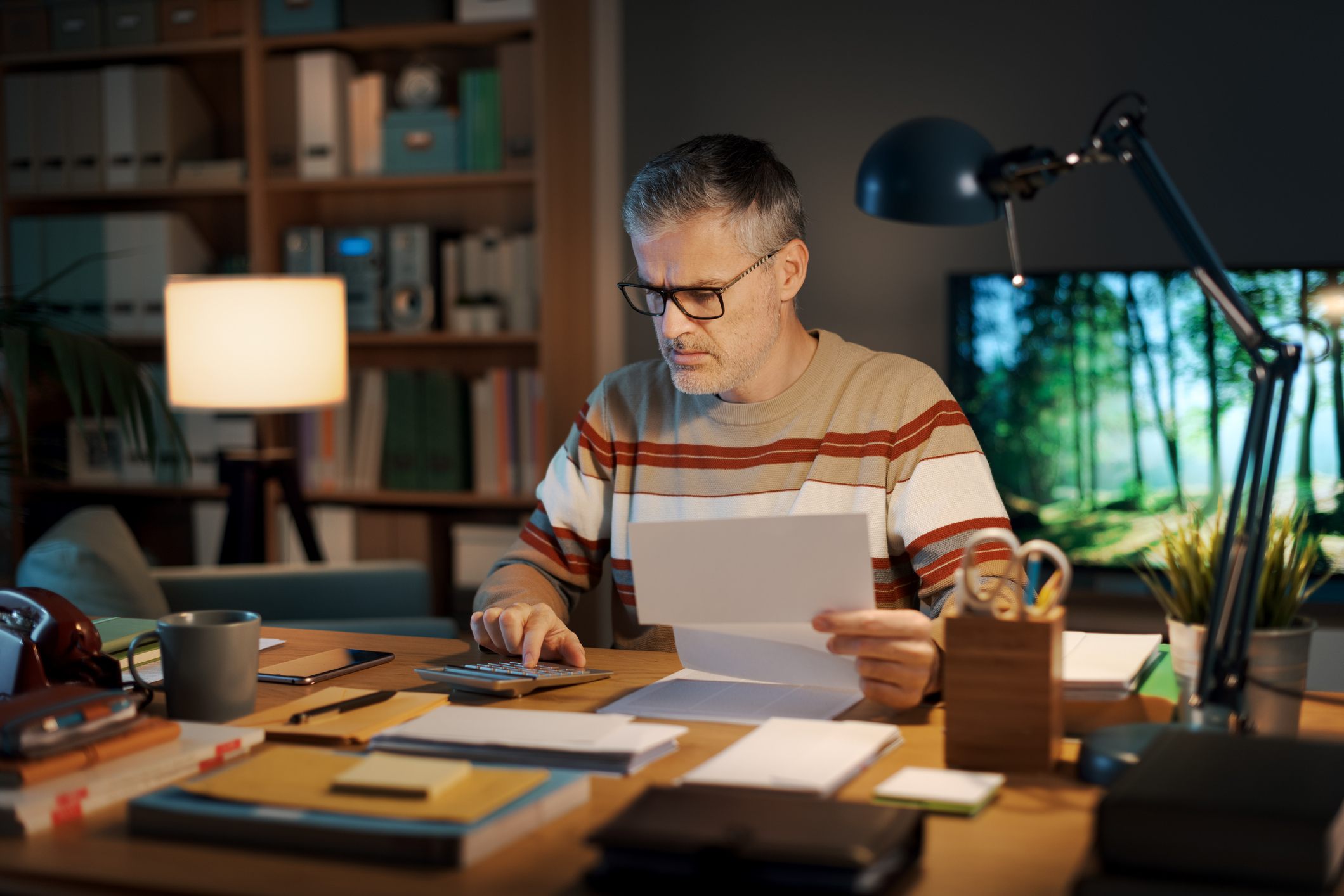 Stressed man sitting at his desk at home and checking an expensive invoice using a calculator.