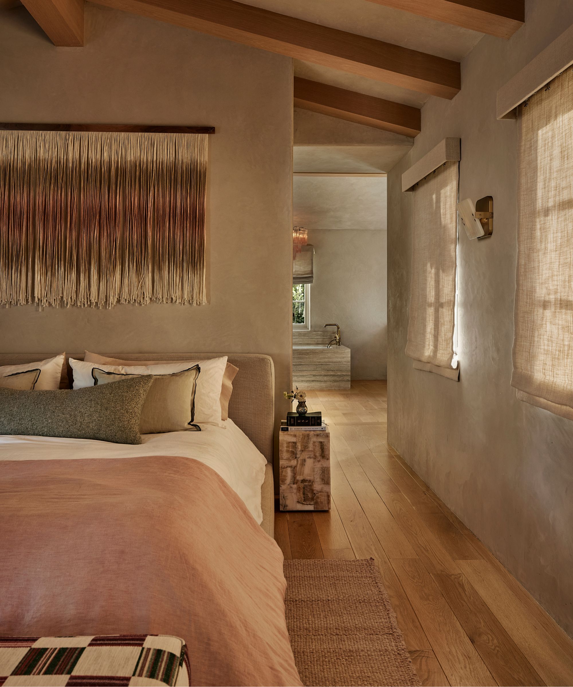 Bedroom with warm taupe plaster walls, exposed wood ceiling beams, linen Roman blinds, and layered neutral bedding, creating a calm, organic retreat with natural textures.