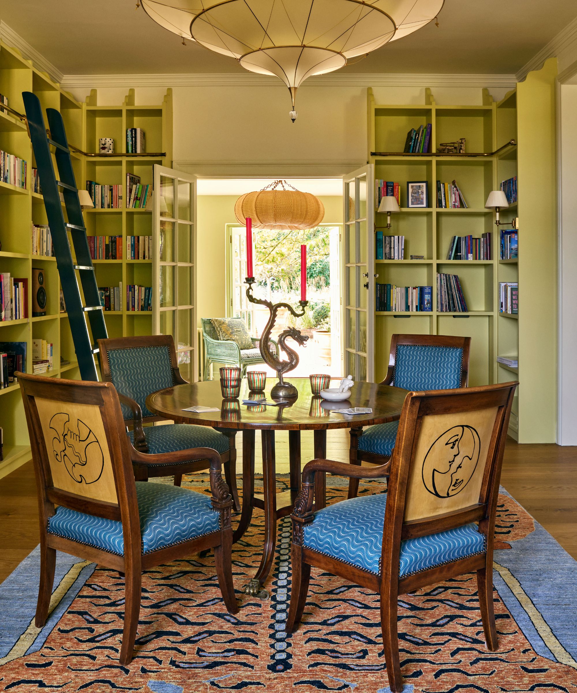 Library with lime green painted built-in bookshelves across the walls, a mahogany round table and blue dining chairs, a salamander rug, a dark green ladder, and a large pendant light.