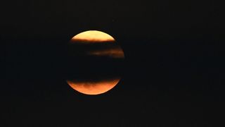 orange hued full moon shining behind a band of black cloud that runs across the center of the moon horizontally.