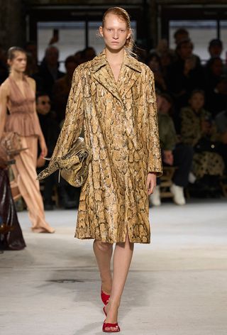 A snakeskin purse is shown in a photo of a model walking down the Dries Van Noten spring 2025 runway show carrying a python handbags styled with a matching trench coat and red mules