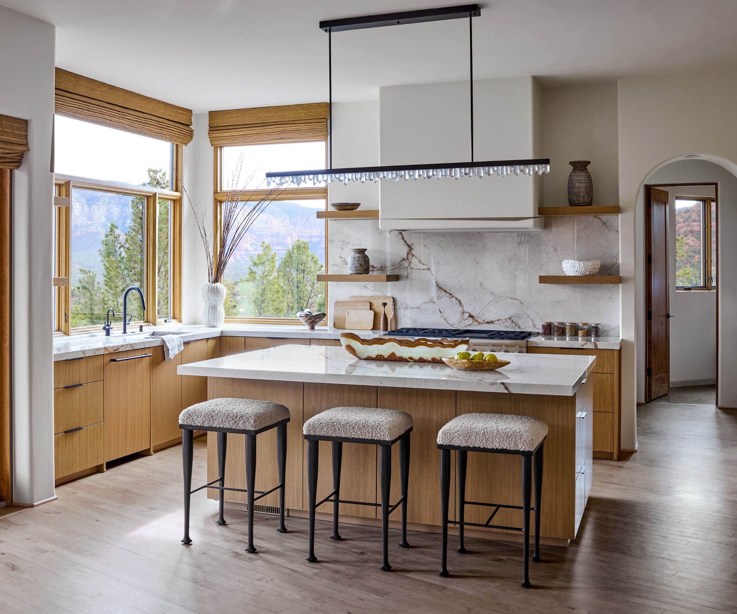 Modern kitchen with wooden island and cabinetry, marble countertops, dakr wood floor and stool surround island, crystal pendant light hanging above island