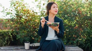 Woman holding up salad and eating sitting on a park bench at lunchtime with a coffee
