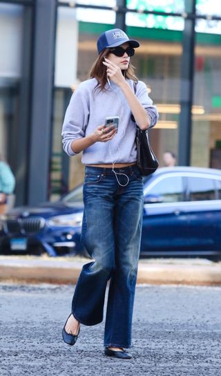 Kaia wears a blue baseball cap, crewneck sweatshirt, blue jeans, black ballet flats, and a black shoulder bag.