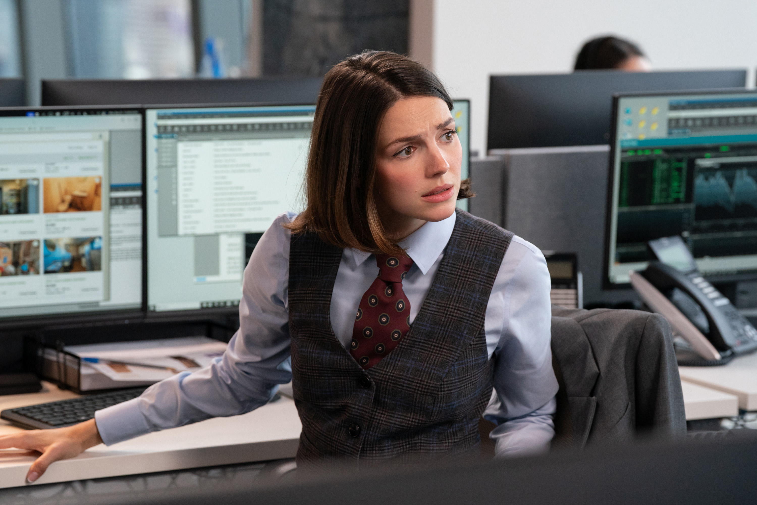 ELLA HUNT as aj wearing a vest and tie sitting at her desk at a finance job in not suitable for work