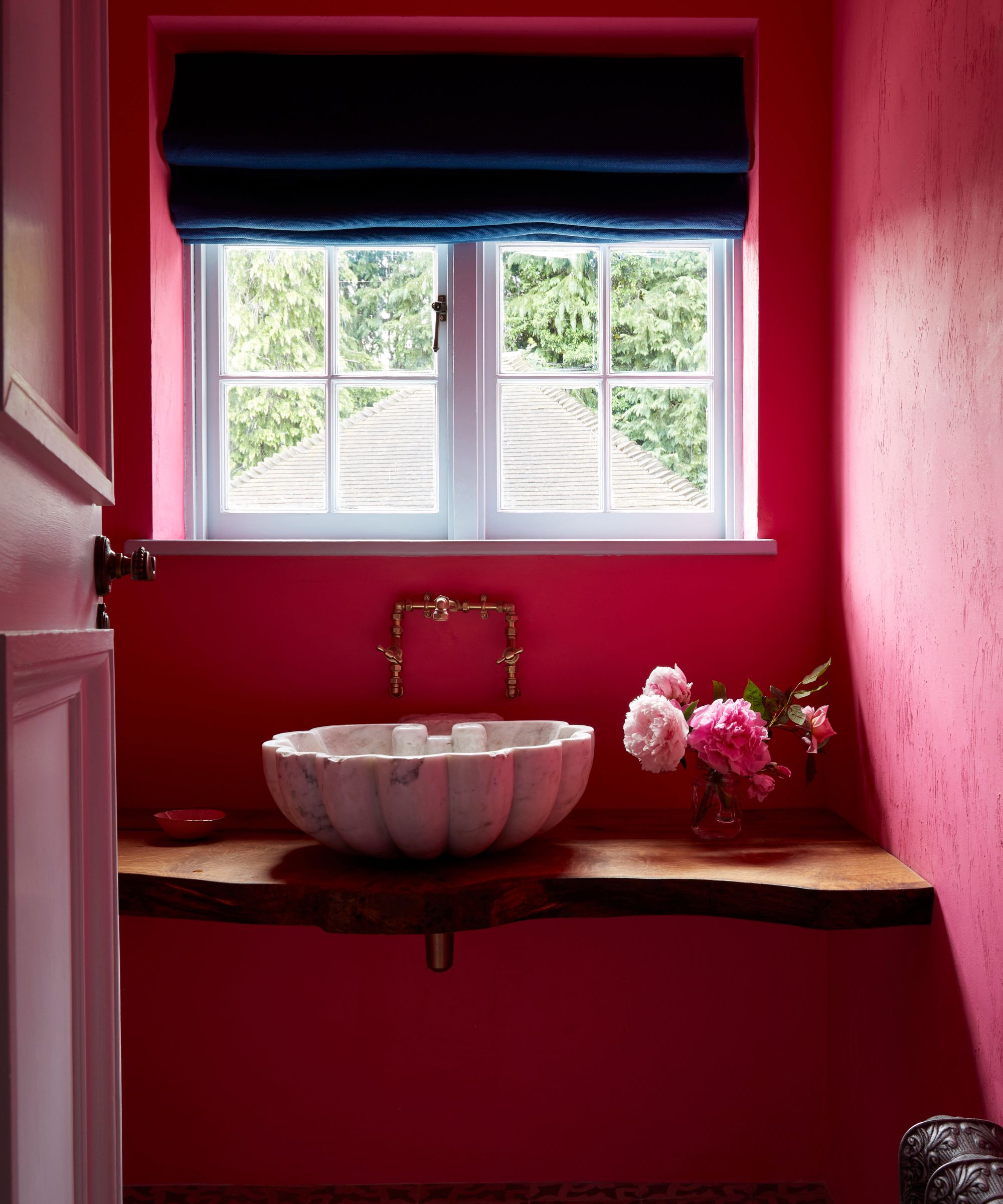 Bathroom with bright pink walls, wooden pedestal with a marble scalloped sink, gold tap, vase of peonies, and a window above with a blue roman blind.