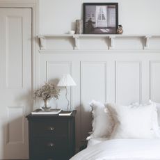 A bedroom with white bedding and cream wall panelling with a contrasting black bedside table displaying a wavy table lamp with a pleated lamp shade