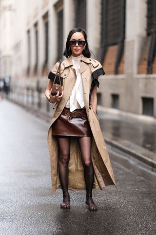 PARIS, FRANCE - MARCH 10: A guest wears black sunglasses, mixed metal rings, white high neck sheer mesh shirt, light brown black trench coat, dark brown suede bag, mixed metal rings, mixed metal bracelets, shiny belted leather mini skirt, black sheer mesh tights, shiny dark brown ballerina flats Maison Margiela Tabi leather shoes, outside Sacai, during the Paris Fashion week Women's Fall/Winter 2025-2026 on March 10, 2025 in Paris, France. (Photo by Edward Berthelot/Getty Images)