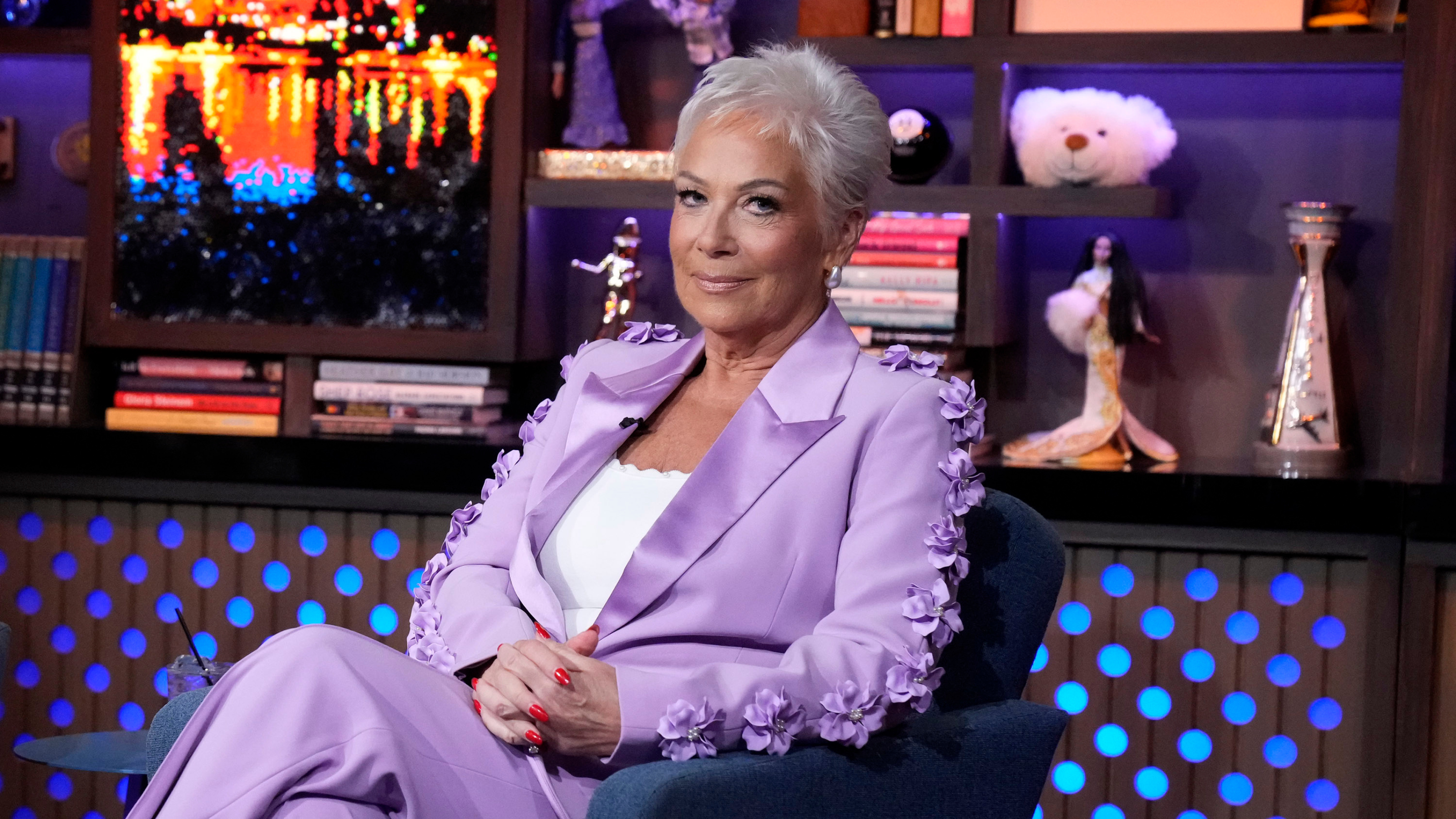 WATCH WHAT HAPPENS LIVE WITH ANDY COHEN -- Episode 22122. Denise Welch on set in lilac, floral suit, sitting on a blue armchair.