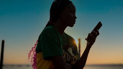 A profile shot of a young woman on a beach using her smartphone, the image is a silhouette as she is standing against a moody sunset