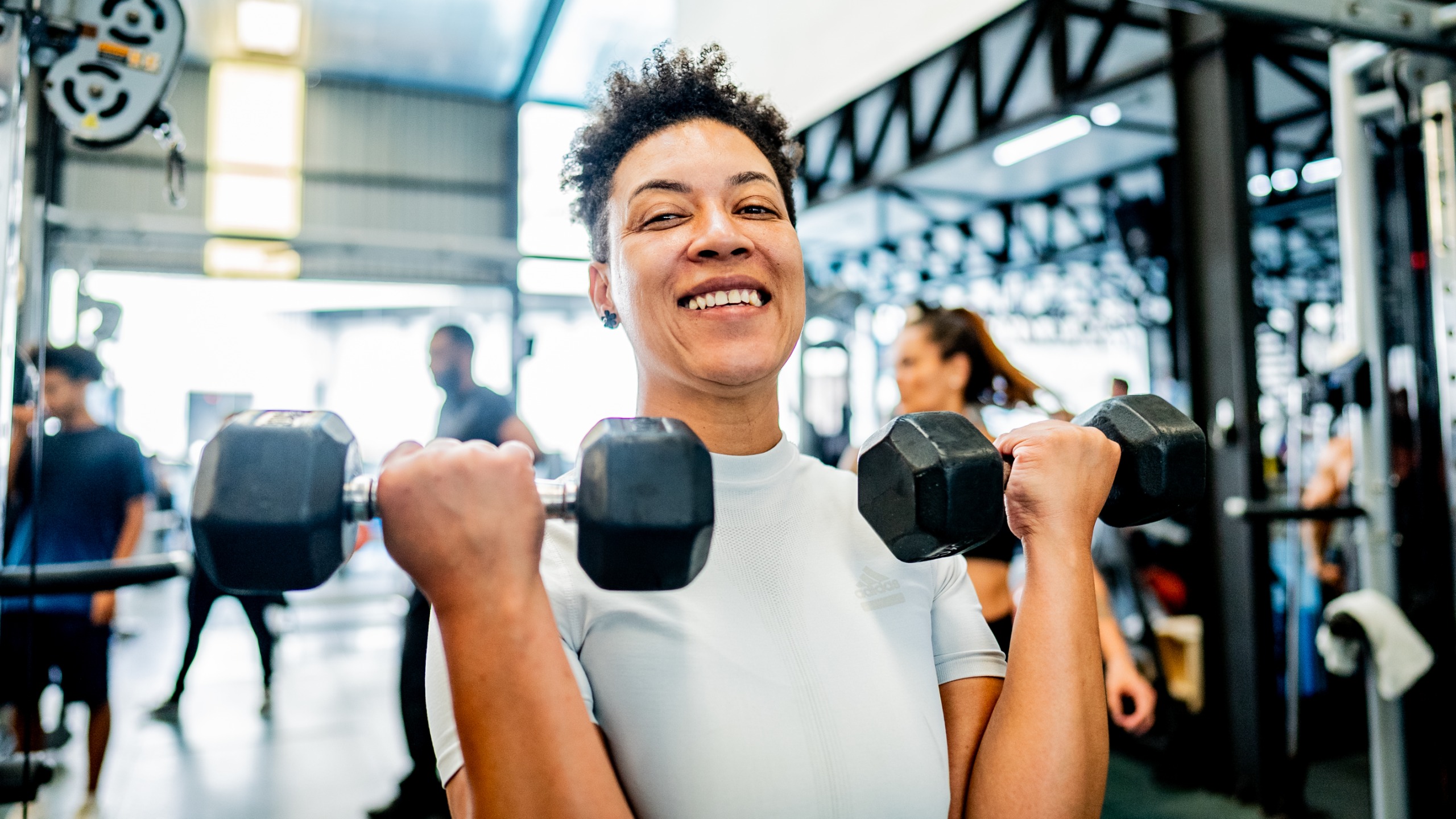A woman in a gym curls two dumbbells up to her shoulders and smiles at the camera. 