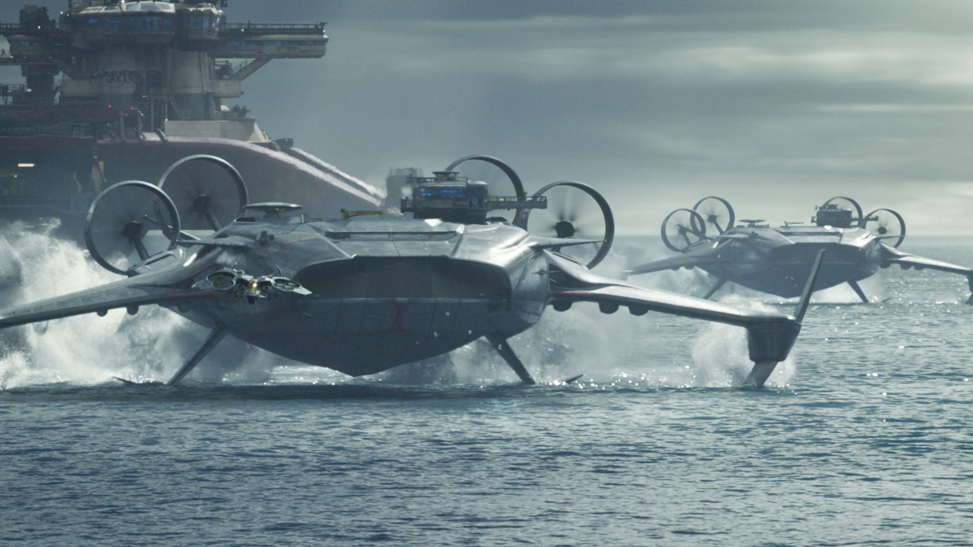 Still from the movie Avatar: Fire and Ash. Two large modern aircrafts are skimming along the water&#039;s surface. Behind them is a giant aircraft carrier-looking vehicle and in front is a small drone.
