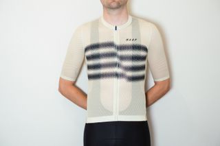 A white man in a pale yellow cycling jersey with black stripes across the chest and black cycling shorts stands against a white wall