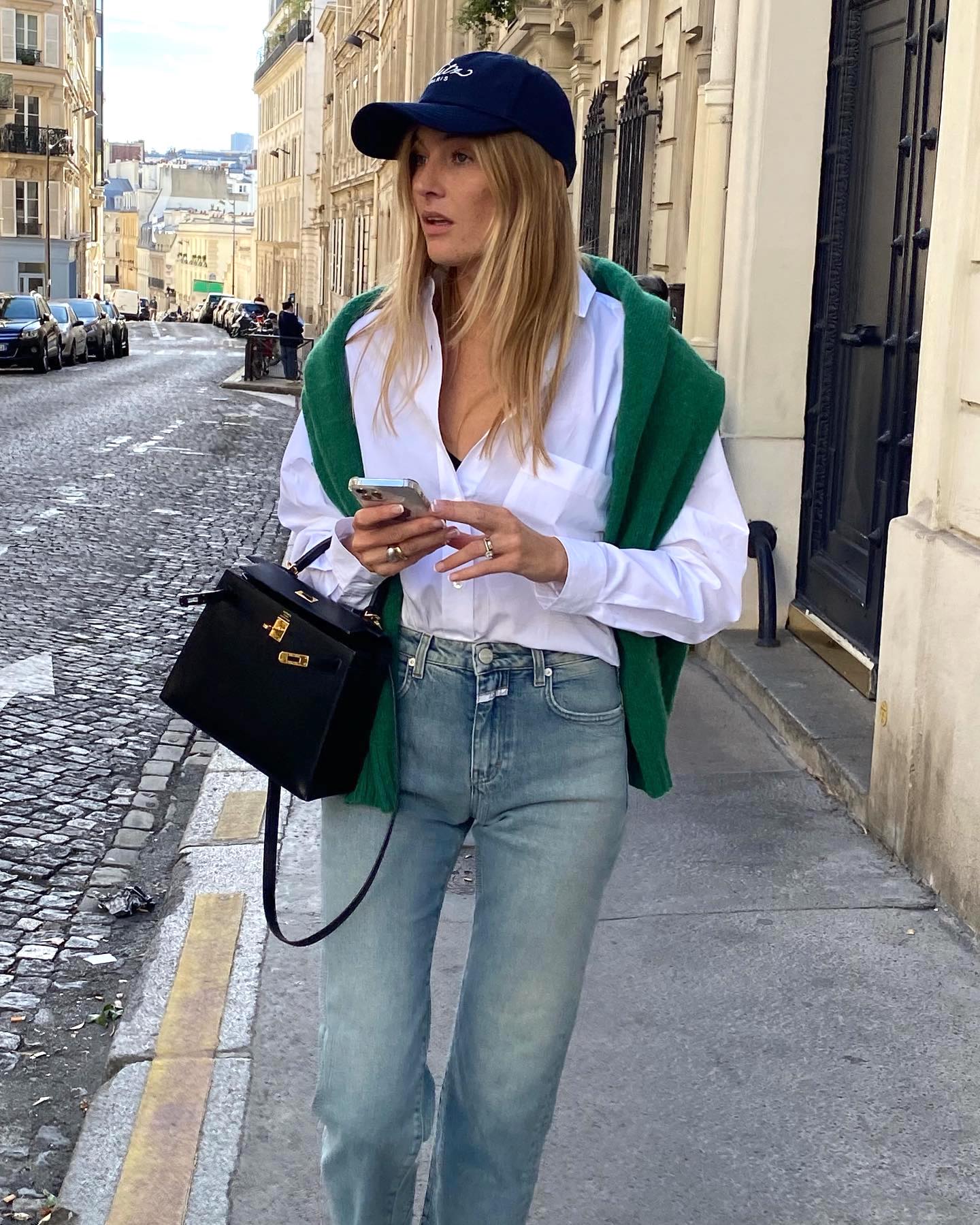 fashion influencer Camille Charriere on a sidewalk in Paris wearing a baseball cap, white button-down shirt, green sweater over the shoulders, jeans, and an Hermès bag