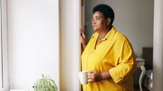 An older woman holds a coffee cup and looks thoughtfully out her bedroom window.