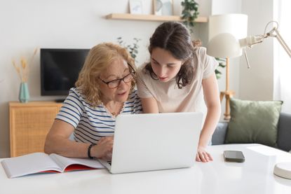 Granddaughter assisting her grandmother with laptop use while sitting together at a table in the cozy living room, fostering connection and learning