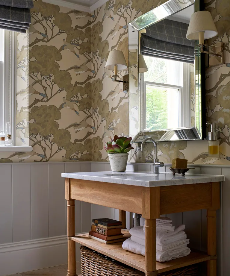 A wallpapered bathroom with a free standing vanity and large mirror. Two sconce lights eitherside of the beveled mirror and towels folded neatly on a shelf of the vanity