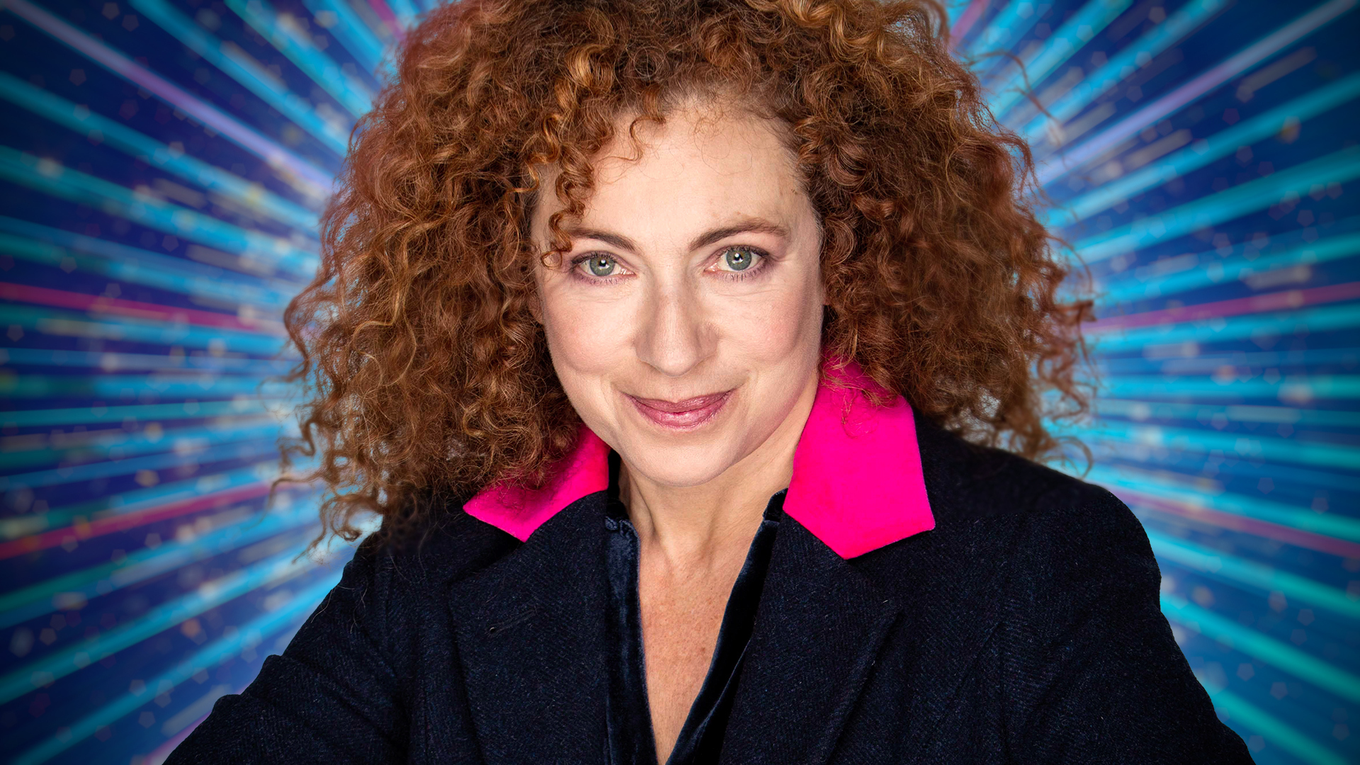 Alex Kingston smiling for the camera with a blue sparkly background behind her.