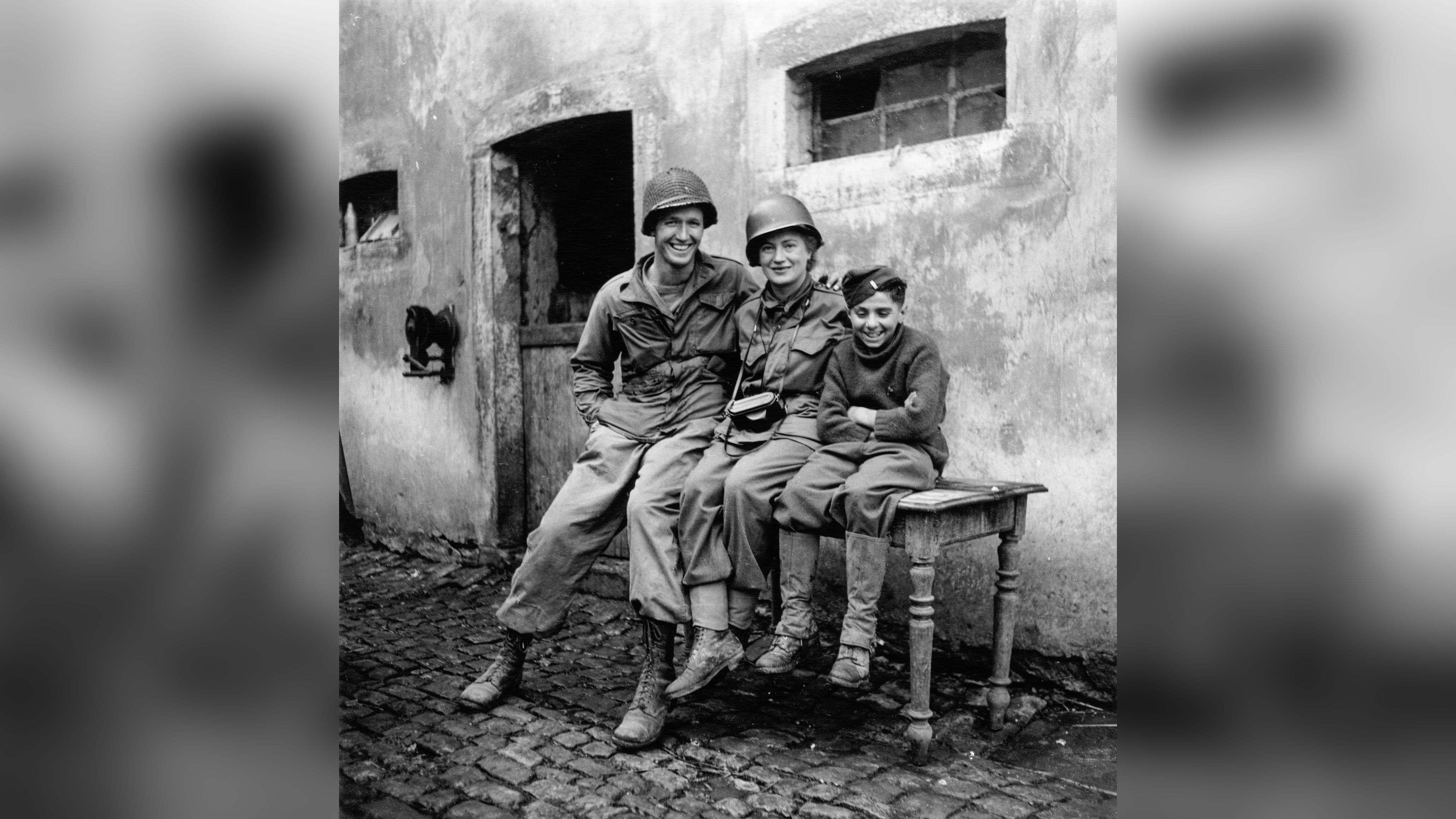 Lee Miller sits grinning between a towering American soldier and a small French boy against a cobbled Luxembourg courtyard wall, her helmet on and camera round her neck. 