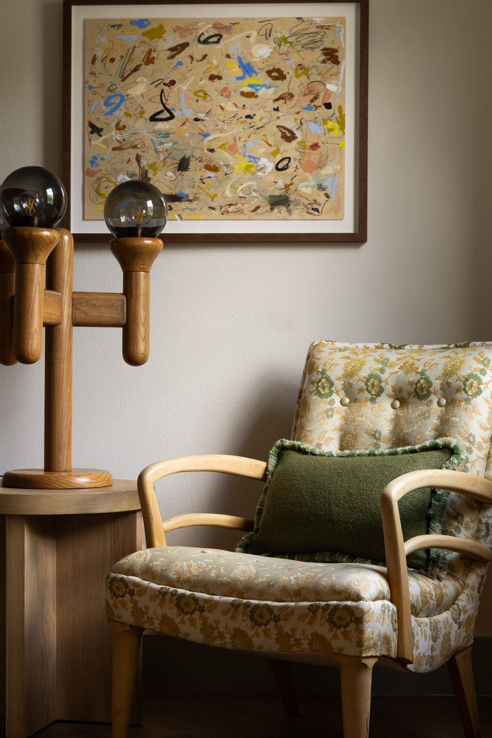 Beige room with colorful abstract art hanging on the wall with a round coffee side table complete with a three pin side lamp and patterned armchair with green cushion