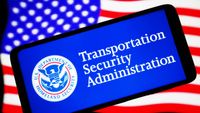 A phone bearing the seal of the US Transportation Security Administration, alongside the words "Transportation Security Administration" in white set against a blue background. The phone is held in front of the USA flag.