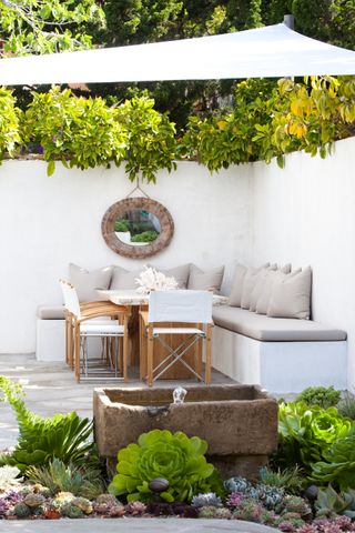 a white walled garden with a white built-in bench, grey cushions and chairs