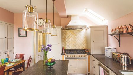 A pink kitchen with marble countertop, gold tiling, and glass and gold pendant lights. To the left is a wooden dining table with burgundy leather chairs.
