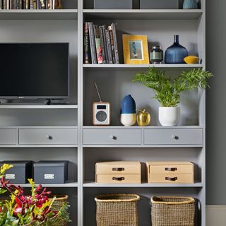 Wall of open shelving in a living room, painted mid grey with space for TV and various storage options