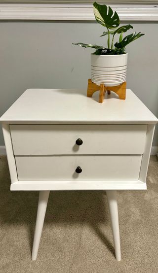 White nightstand with spindly legs and two drawers with round black handles. On top, a white pot plant with a baby monstera plant
