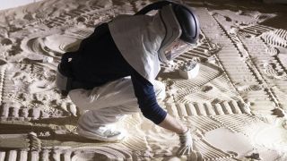 a technician in a respirator kneels in a sandbox full of ridges and mounds