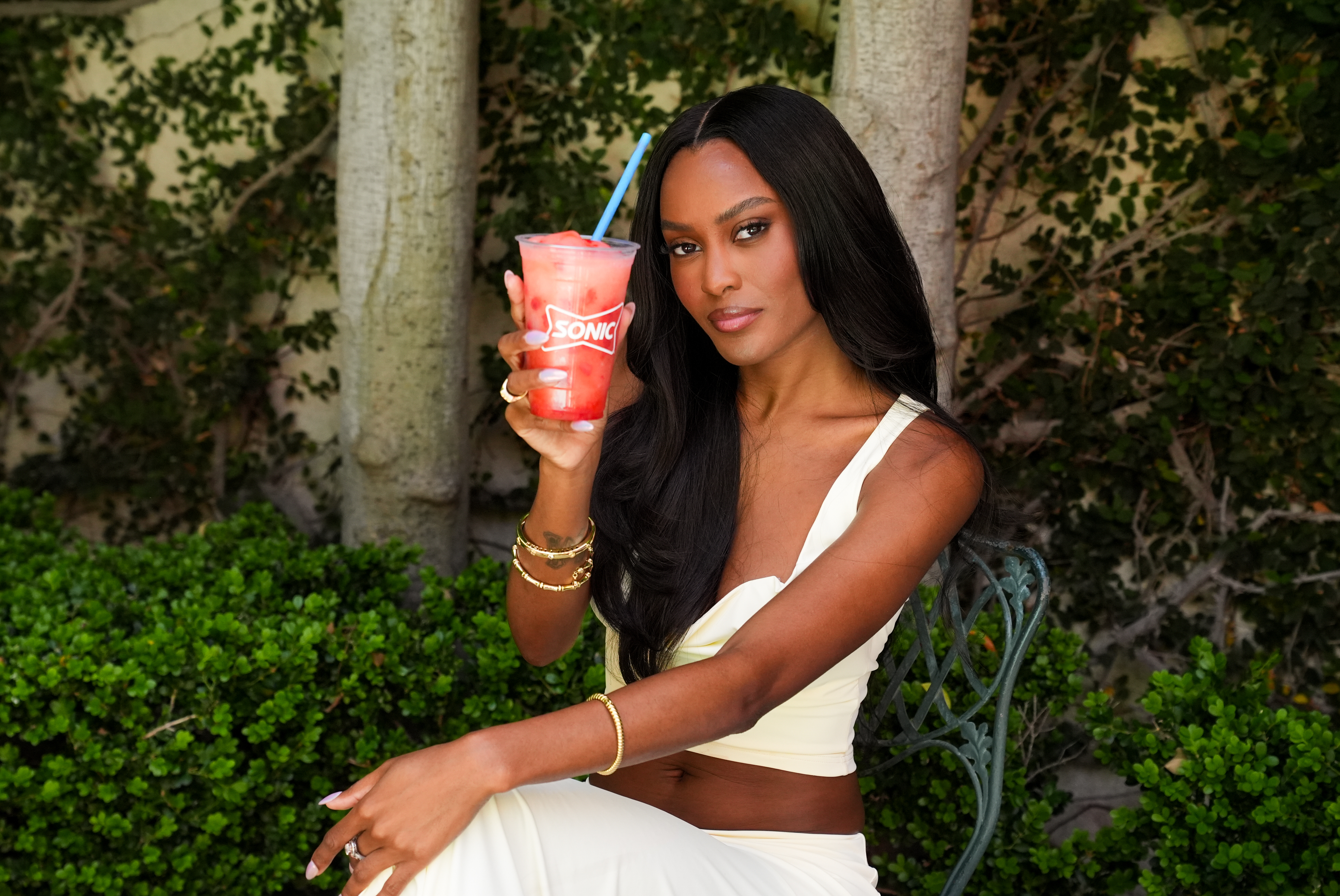 Ciara Miller poses with a Sonic Frozen Refresher while wearing an all-white outfit and sitting in a wire garden chair in front of foliage.