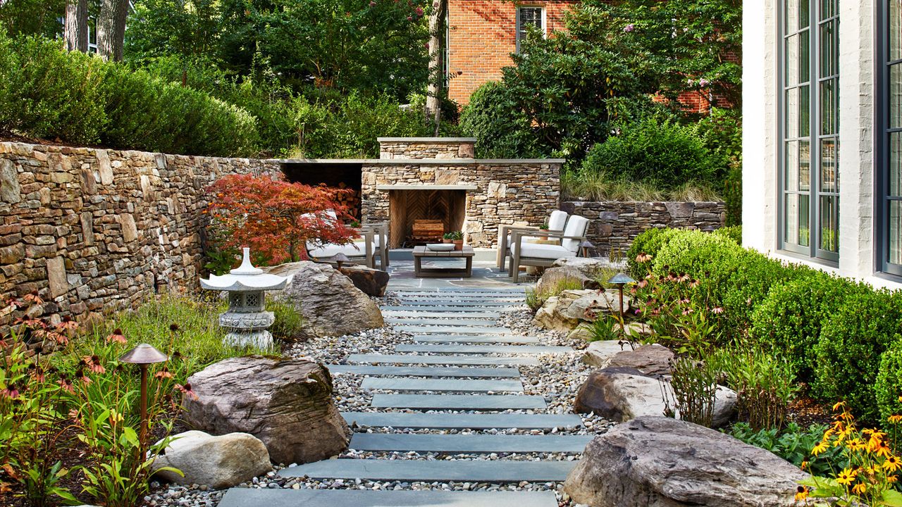 slate and gravel garden path edged with boulders and planting beds including a low hedge, and a brick wall, with an outdoor fireplace and garden furniture