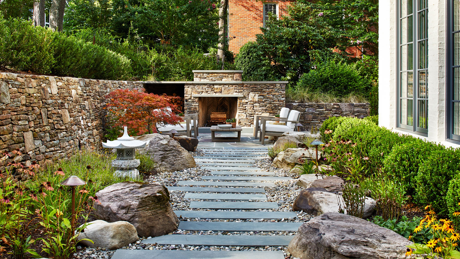 slate and gravel garden path edged with boulders and planting beds including a low hedge, and a brick wall, with an outdoor fireplace and garden furniture