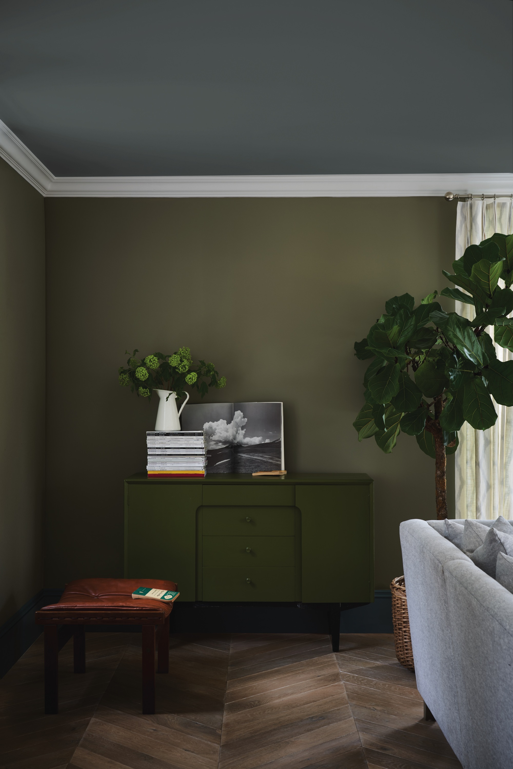 a color blocked green and gray living room with timber parquetry floors, and green console styled with artwork and a stack of books topped with a vase of flowers, a brown leather stool, the back of a gray sofa, and a potted fiddle leaf tree