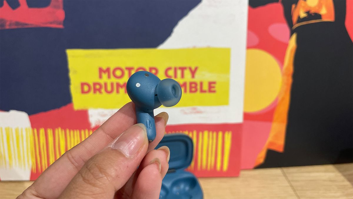 A hand holding one of the JBL Live Beam 3 in-ear headphones. The open case is just visible in the background.