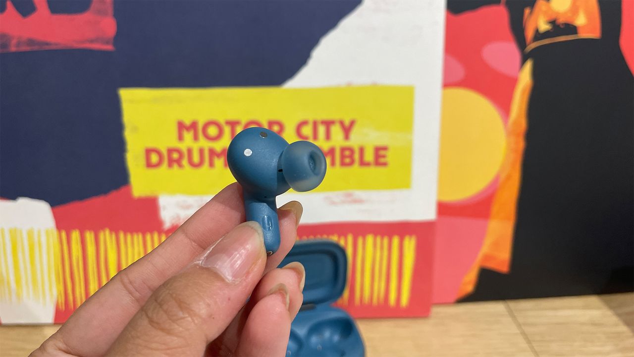 A hand holding one of the JBL Live Beam 3 in-ear headphones. The open case is just visible in the background.