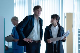 Beyond Paradise season 2 episode 5: DI Humphrey Goodman (Kris Marshall) and DS Esther Williams (Zahra Ahmadi) stand in a hospital ward. He has his hands on his hips and she is holding an open folder of medical notes.