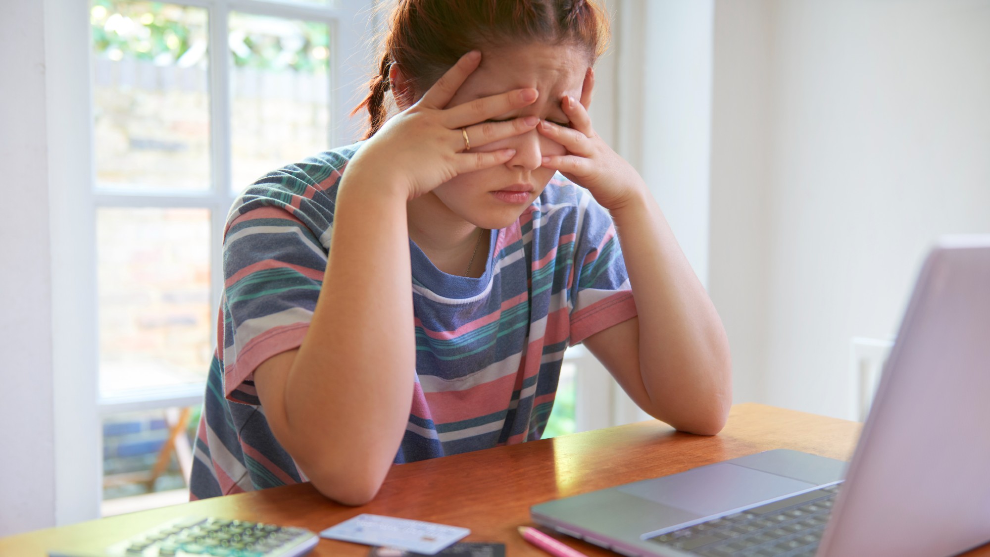 Worried student with her head in her hands sitting at a desk in front of her laptop, calculating student loan debt 