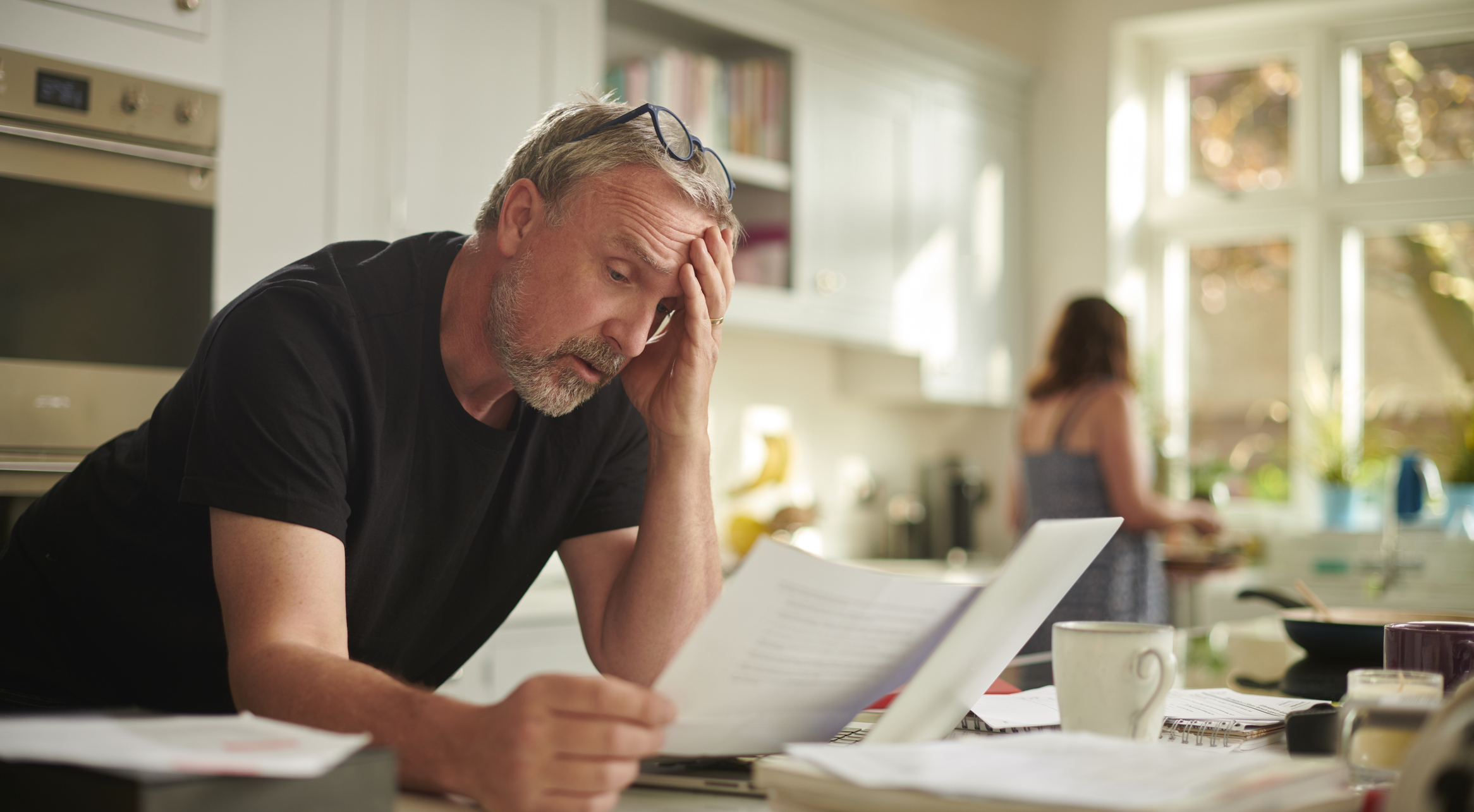 A mature man in his kitchen looking stressed while looking at bills.