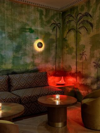 A creatively designed restaurant features a blend of Art Deco, modern glamour, and Space Age design furnishings, including golden sconces, red table lamps, and golden coffee tables, all of which contrast with its tropical-inspired, palm tree-filled wallpaper.